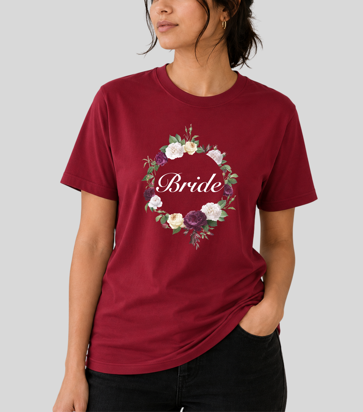 A woman wearing a burgundy short-sleeve t-shirt with a floral wreath graphic and the word 'Bride' in white script across the chest, paired with black jeans, against a solid gray background.