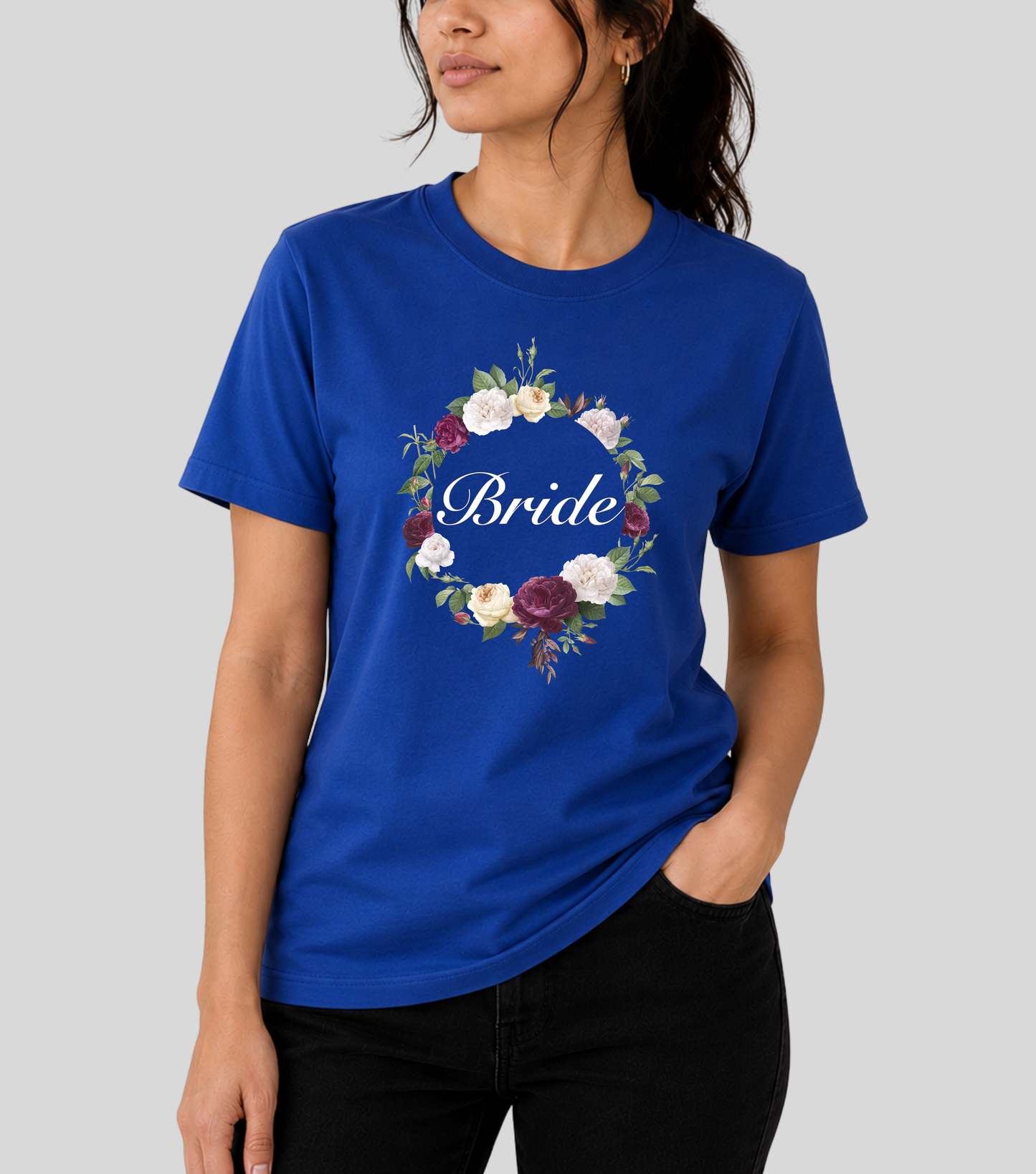 A woman wearing a royal blue crewneck t-shirt with a floral wreath graphic and the word 'Bride' in white script across the chest, paired with black pants, against a solid gray background.