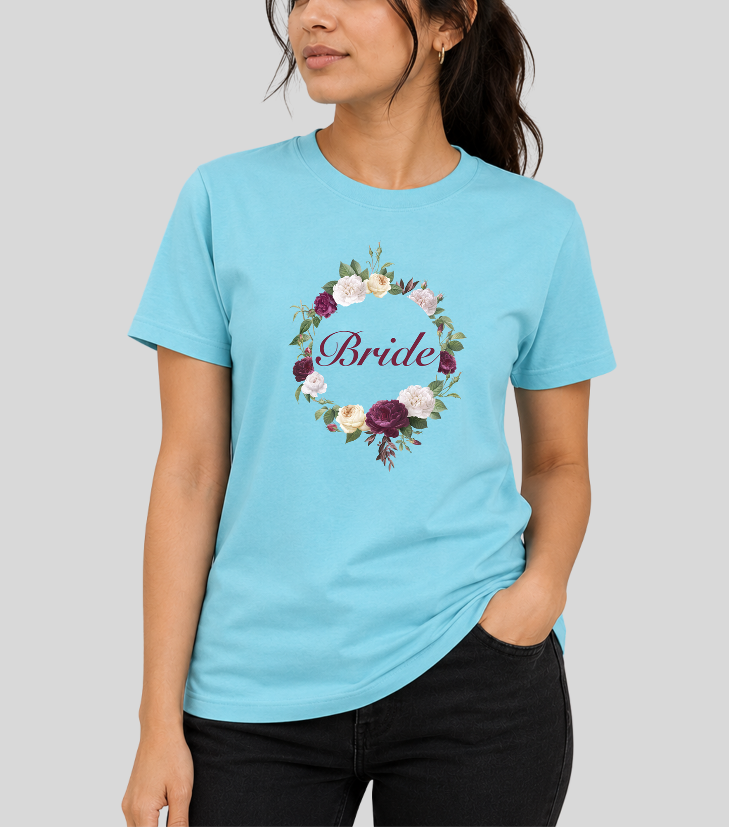 A woman wearing a light blue crewneck t-shirt with a circular floral wreath graphic on the chest containing the word 'Bride' in purple script. The background is a solid light gray studio backdrop.