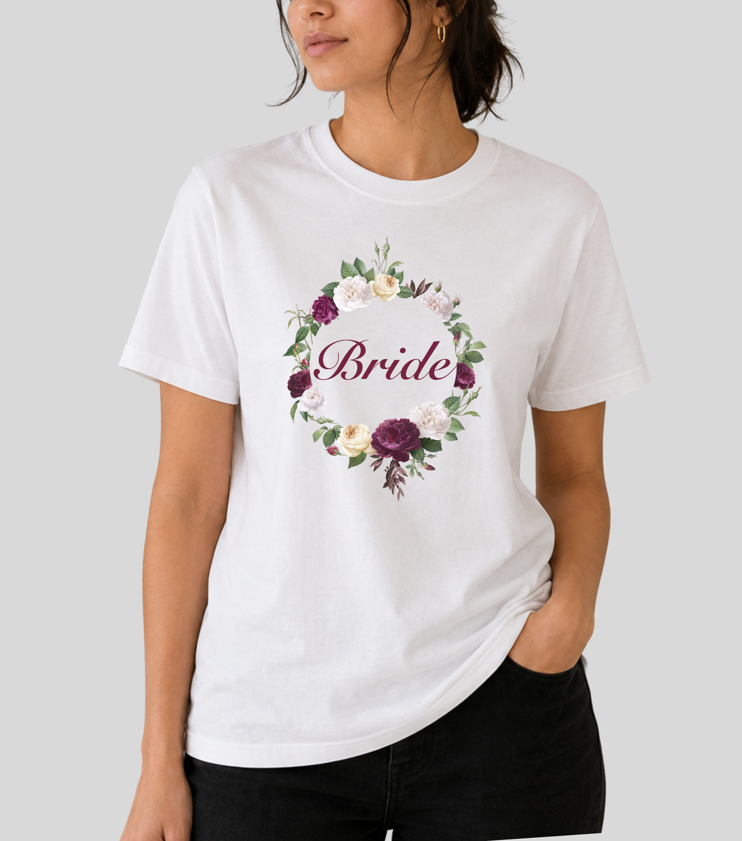 A woman wearing a white short-sleeve crewneck t-shirt with a floral wreath graphic and the word 'Bride' in script across the chest, paired with black pants, against a solid light gray background.