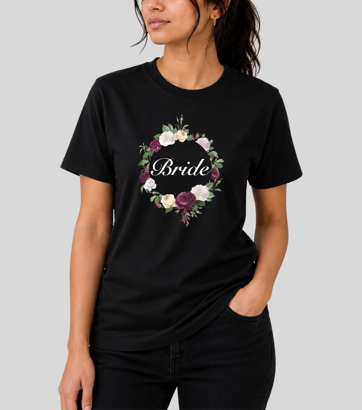 A woman wearing a black short-sleeve crewneck t-shirt with a floral wreath graphic and the word 'Bride' in script across the chest, paired with black jeans, against a solid gray background.
